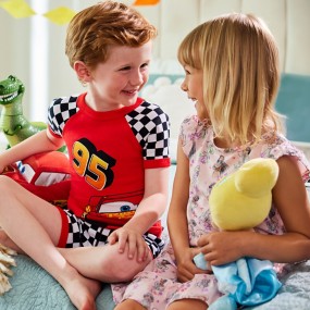Young boy with red hair wearing Lightning McQueen pajamas sitting on a bed with a young blonde girl wearing a Disney princess nightgown with toy story action figures in the background.