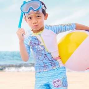 Boy wearing Star Wars-themed swimwear stands on beach with beach ball, blue snorkeling mask, cheeks puffed out.