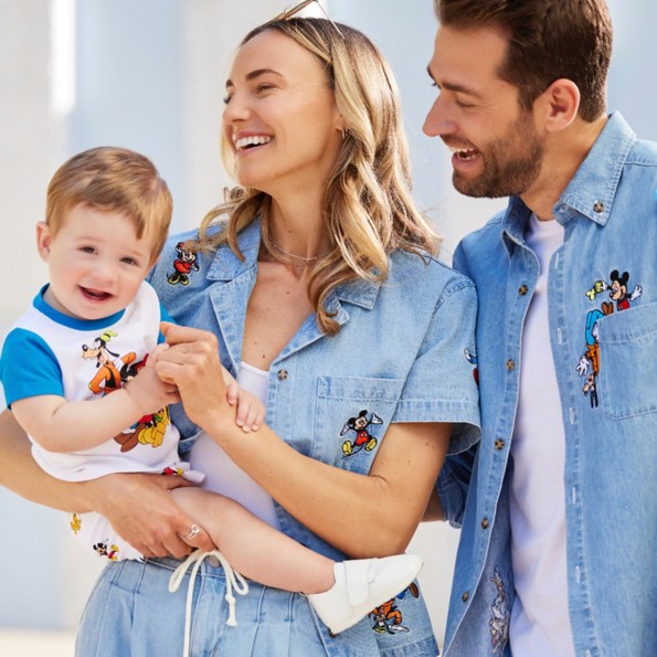 Family of three in matching outfits. Mom holds toddler boy in Mickey & Friends tshirt and shorts. Mom wears denim shirt with Mickey and Minnie embroidery. Dad wears denim shirt with Mickey and Goofy embroidery.