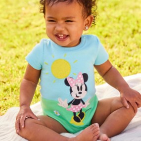 Baby girl sitting on a blanket outside wearing a Minnie Mouse onesie featuring Minnie in a pink dress on a sunny day.
