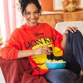 Brunette woman sitting on a couch eating popcorn wearing dark gray sweatpants and an orange Lion King Celebration Crew long sleeve shirt.