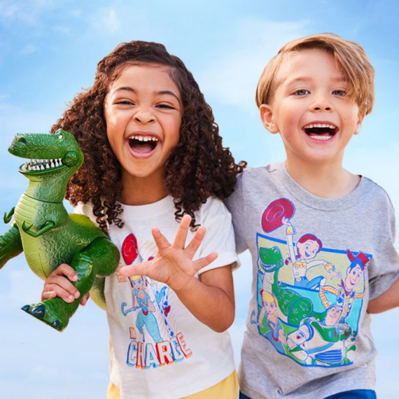 Young brunette girl holding a Rex action figure wearing a white, red and blue Toy Story t-shirt standing next to a blonde young boy holding a Buzz Lightyear action figure wearing a gray, blue and red Toy Story t-shirt.