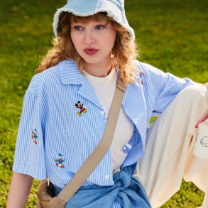Woman in blue & white seersucker button down with Mickey & Friends embroidery, tan waist bag, denim bucket hat sitting outside.