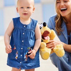 Toddler girl in denim dress with Mickey & Friends details, mom holding Pluto plush behind her.