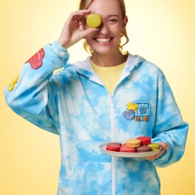 Blonde woman wearing Walt Disney World Blue Tie-Dye Zip-Up Hoodie holding a plate of macarons and one over her eye.
