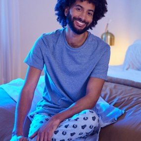 A happy man sitting on a bed wearing a grey t-shirt and Star Wars pajama pants with Darth Vader helmets and Empire symbol details.
