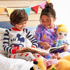 Young boy with red hair wearing Lightning McQueen pajamas sitting on a bed with a young blonde girl wearing a Disney princess nightgown with toy story action figures in the background.