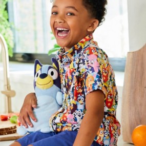 Young boy with brown curly hair sitting on a counter wearing a Bluey patterned RSVLTS button-down shirt featuring a repeating pattern of the Bluey characters.
