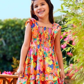 Young girl with brown hair wearing an Encanto-themed vibrant coral peasant dress for girls with colorful florals and butterflies in blue, pink, yellow, and green.